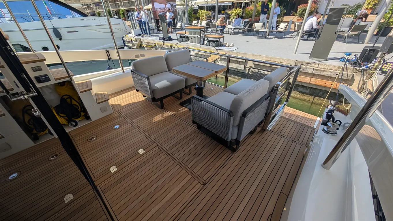 Boating deck featuring two grey armchairs and a wooden table, overlooking a marina with people and boats in the background.